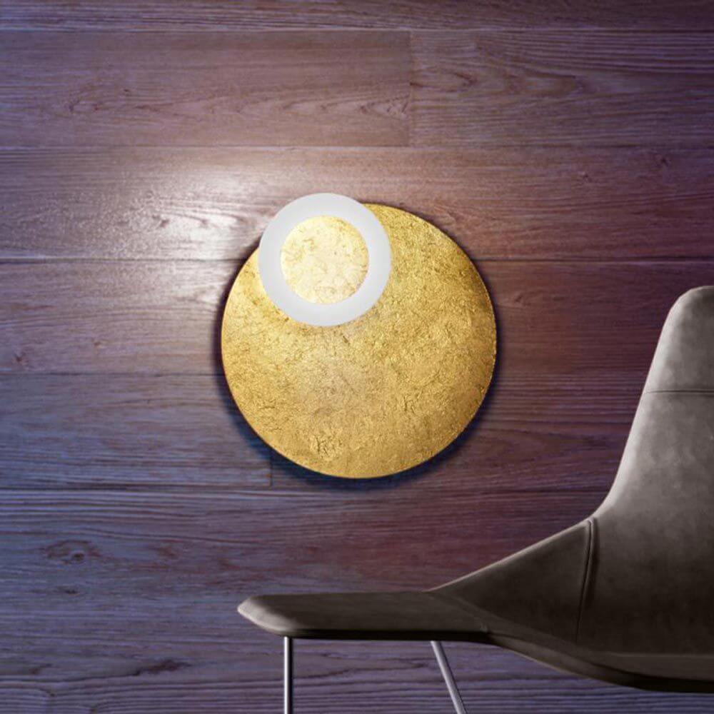 Moderne Icone LED Deckenleuchte Vera in Gold und Weiß, Ø 50cm, an Wand, mit stilvollem Stuhl daneben.