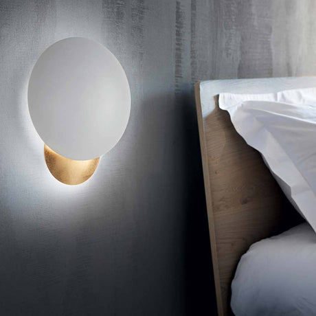 Eclissi LED Væglampe hvid, guld på en væg ved siden af en seng, stilfuld belysning til moderne rum.