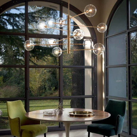 Suspension Equinoxe à 12 flammes en laiton antique dans un espace repas moderne avec de grandes fenêtres et des chaises élégantes.