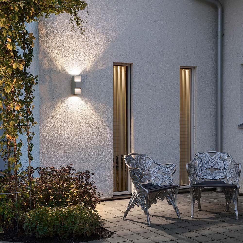 Imola LED Außen-Wandleuchte Style Design Grau, opales Acrylglas an einer Hauswand mit Gartensitzmöbeln.
