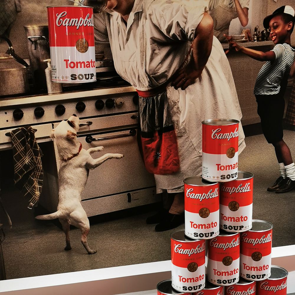 Ein Stapel Campbell's Tomatensuppe-Dosen in einer Küche mit einem Hund und Koch, die zusammenarbeiten.