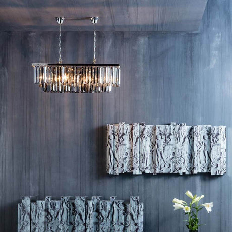 Hängeleuchte Smoky Lounge Rectan mit facettierten Glaskristallen über Wandrelief und Blumenarrangement.