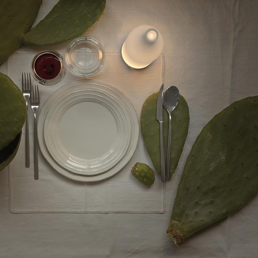 Karman Bacco LED-Tischleuchte in Flaschenform auf einem gedeckten Tisch mit Kakteen.