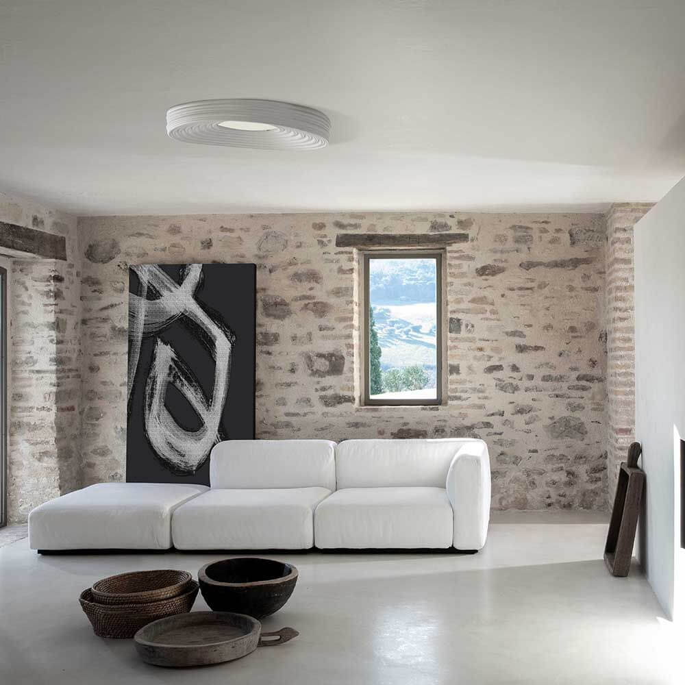 Moderne Wohnlandschaft mit Karman LED-Deckenleuchte R.O.M.A. über einem weißen Sofa in rustikalem Raum.