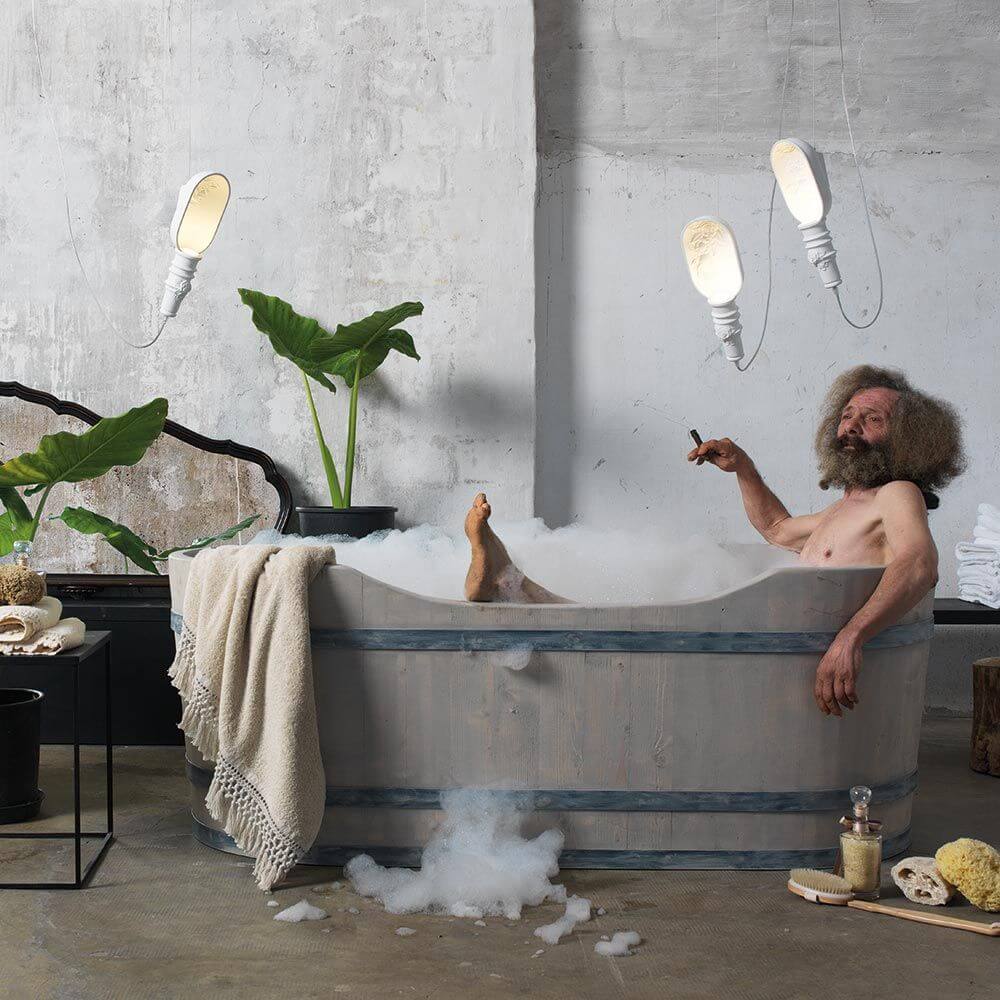Mann entspannt in einer Badewanne mit Karman Workinprogress Hängeleuchte Weiß im Hintergrund, umgeben von Pflanzen.