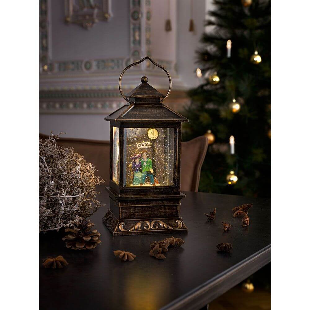 LED Schneelaterne mit Pärchen klein Timer wassergefüllt batteriebetrieben auf einem Tisch mit Tannenzapfen und Weihnachtsbaum im Hintergrund.