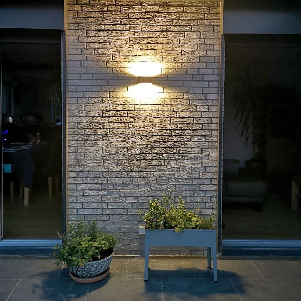 LED Außenwandleuchte Gemini IP54 in Anthrazit an einer Steinwand mit Pflanzen im Vordergrund.