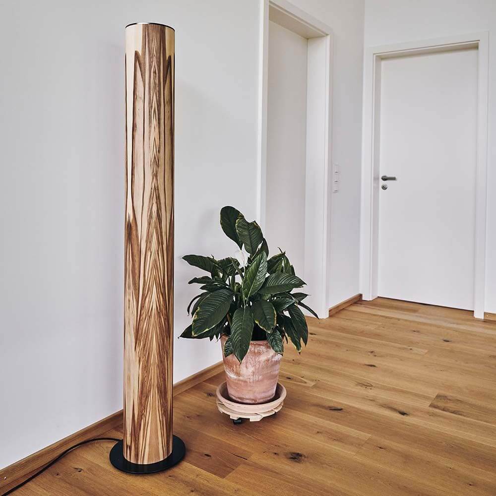 LED Holz-Stehleuchte Lucerna 160cm Satin Nussbaum steht neben einer Pflanze in einem modernen Raum.