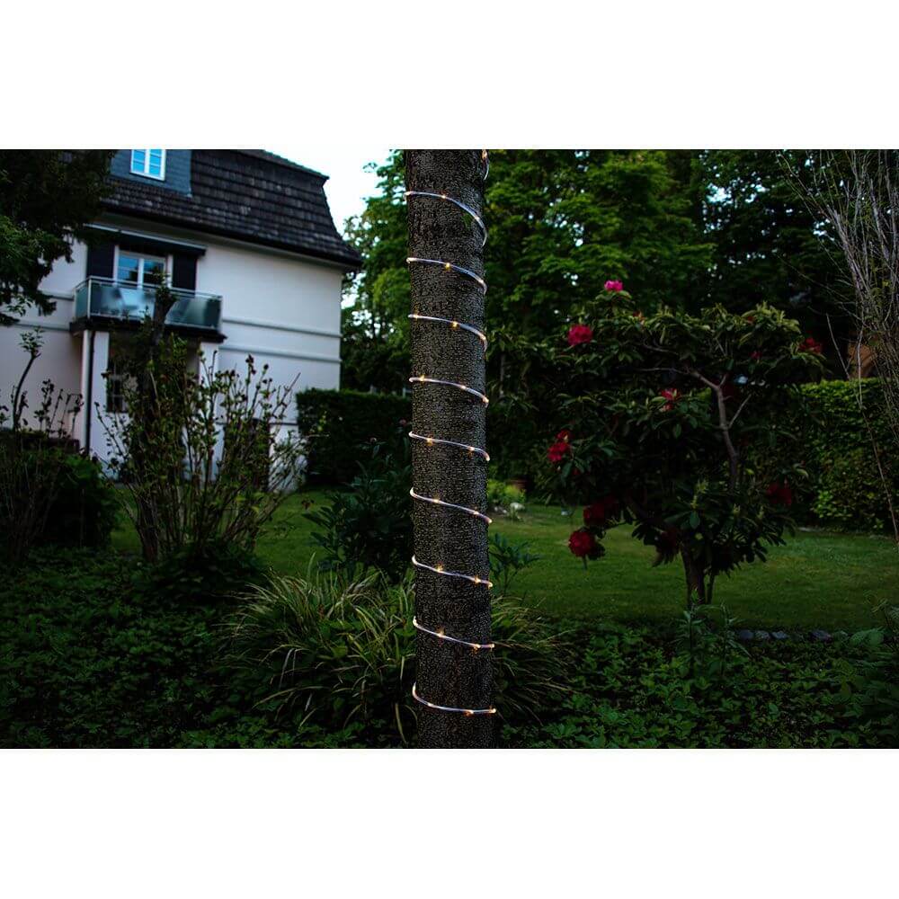 LED Mini Lichterschlauch 10m mit Lichtsensor Timer 130 Warmweiße Dioden batteriebetrieben IP44 um einen Baum im Garten gewickelt.