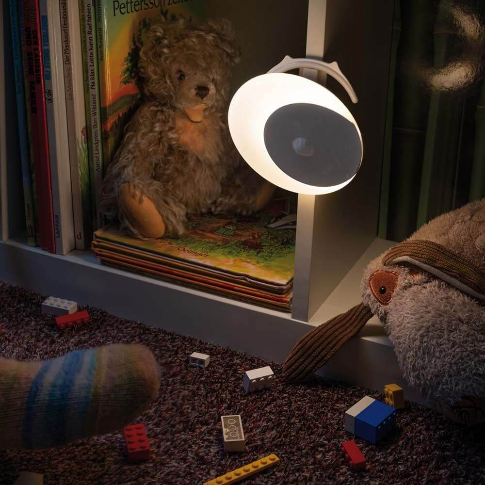 LED Nachtlicht Viby mit Dämmerungssensor leuchtet sanft neben Spielzeug und Bücherregal in dunklem Raum.