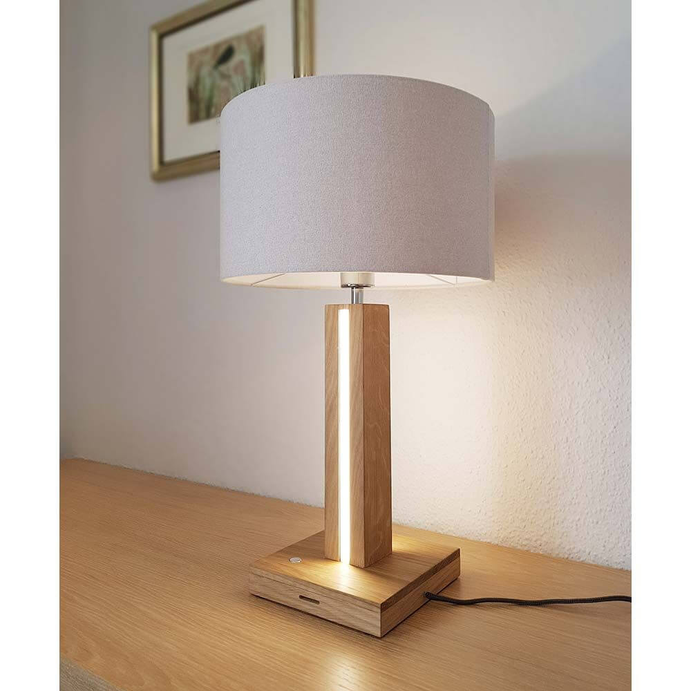LED Tischlampe Asseto 980lm Eiche-Geölt, Beige auf einem Holztisch mit grauem Lampenschirm und sanftem Licht.