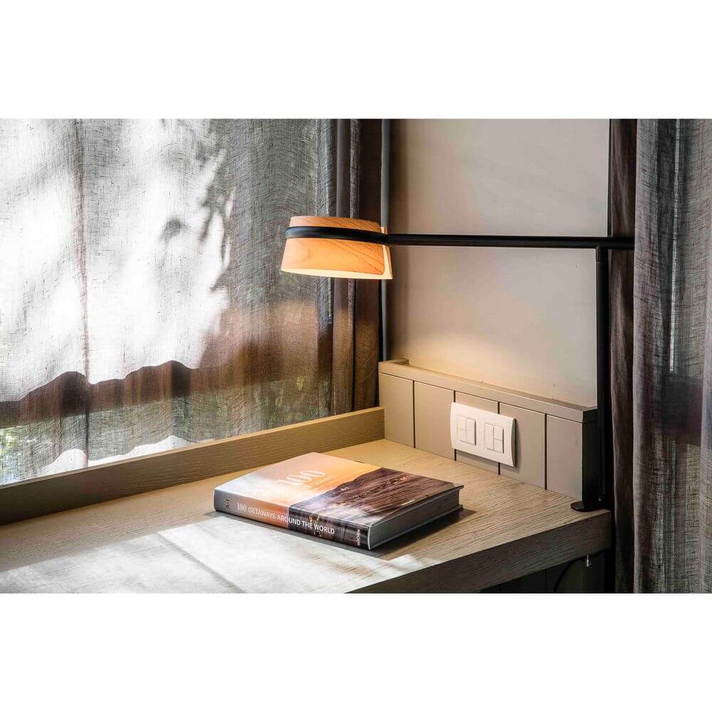 LED Tischlampe LOOP Schwarz, Braun auf einem Schreibtisch mit einem Buch und Vorhängen im Hintergrund.