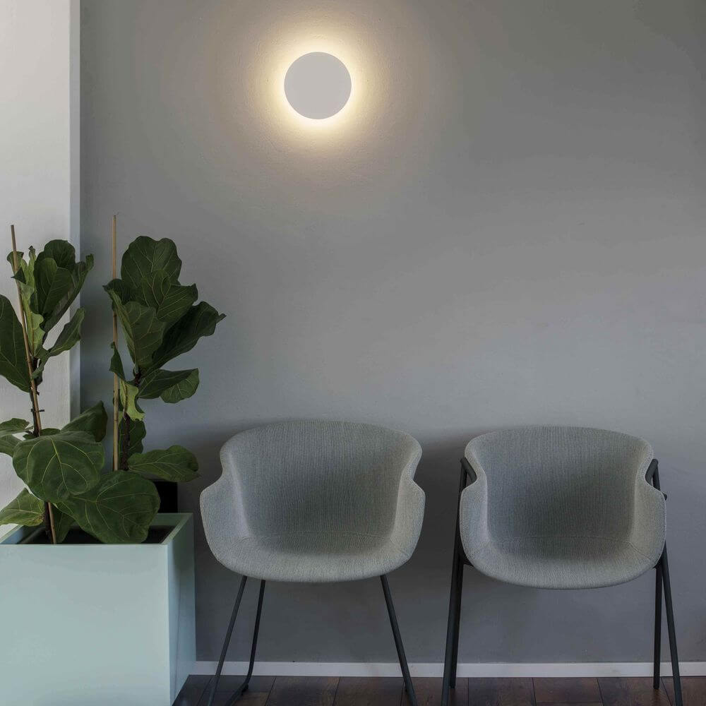 Moderne LED Wandleuchte LUNA 5W 2700K Weiß an der Wand über zwei grauen Stühlen in einem gemütlichen Raum.