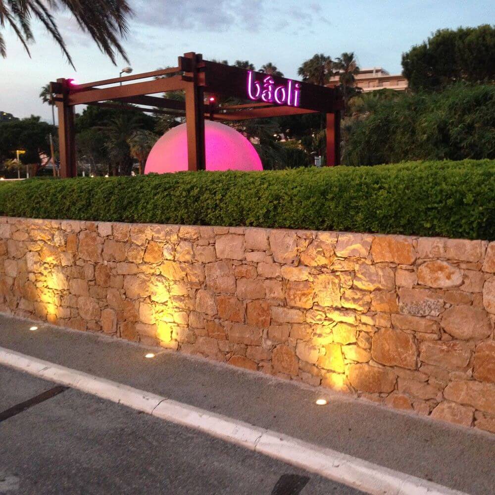 Baoli Restaurant mit Leuchtschrift in der Abenddämmerung, umgeben von grünen Hecken und beleuchtetem Steinmauerwerk.