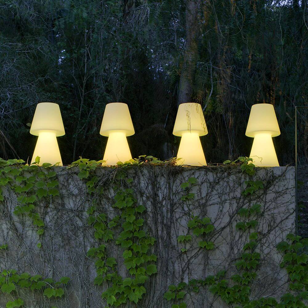 LED-Akku-Tischleuchte Lola Large mit Fernbedienung in modernem Design auf einer Mauer mit Pflanzen.