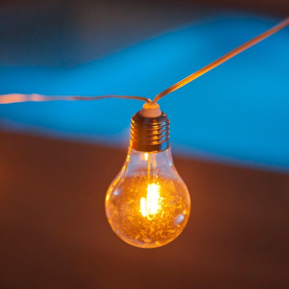 Nahaufnahme einer warmen Glühbirne in einer Lichterkette über einem Pool bei Nacht.