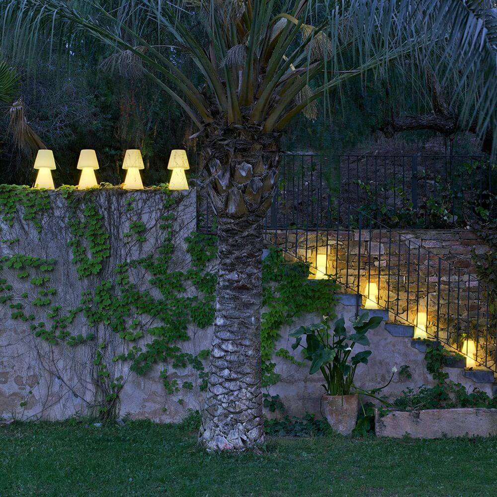 Lola LED-Außen-Tischleuchte Large in einem Garten, umgeben von Pflanzen und stimmungsvollem Licht.