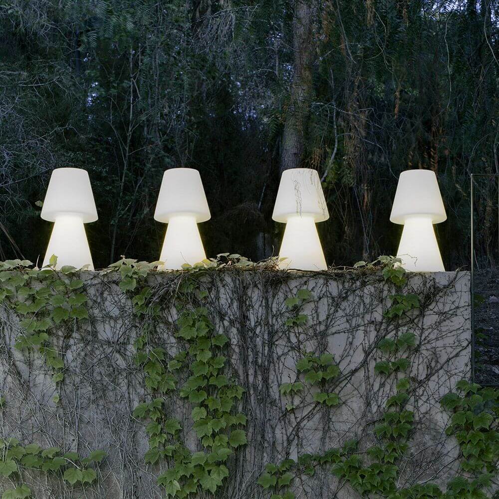 Lola LED-Außen-Tischleuchte Large in einer Gartenumgebung, vier stilvolle Leuchten auf einer Mauer umgeben von Pflanzen.