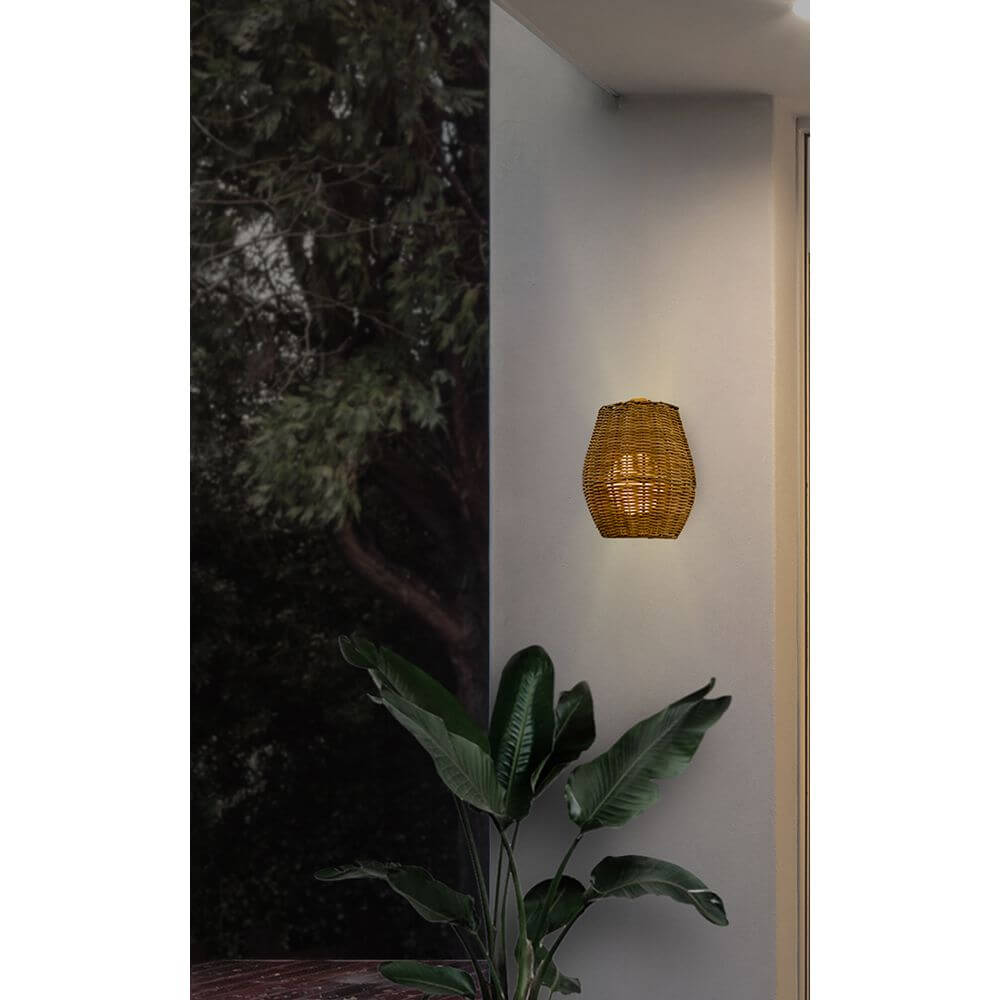 Saona Boho-Style LED Wand-Akkuleuchte aus Rattan IP54 an einer Wand mit Pflanzen im Vordergrund.