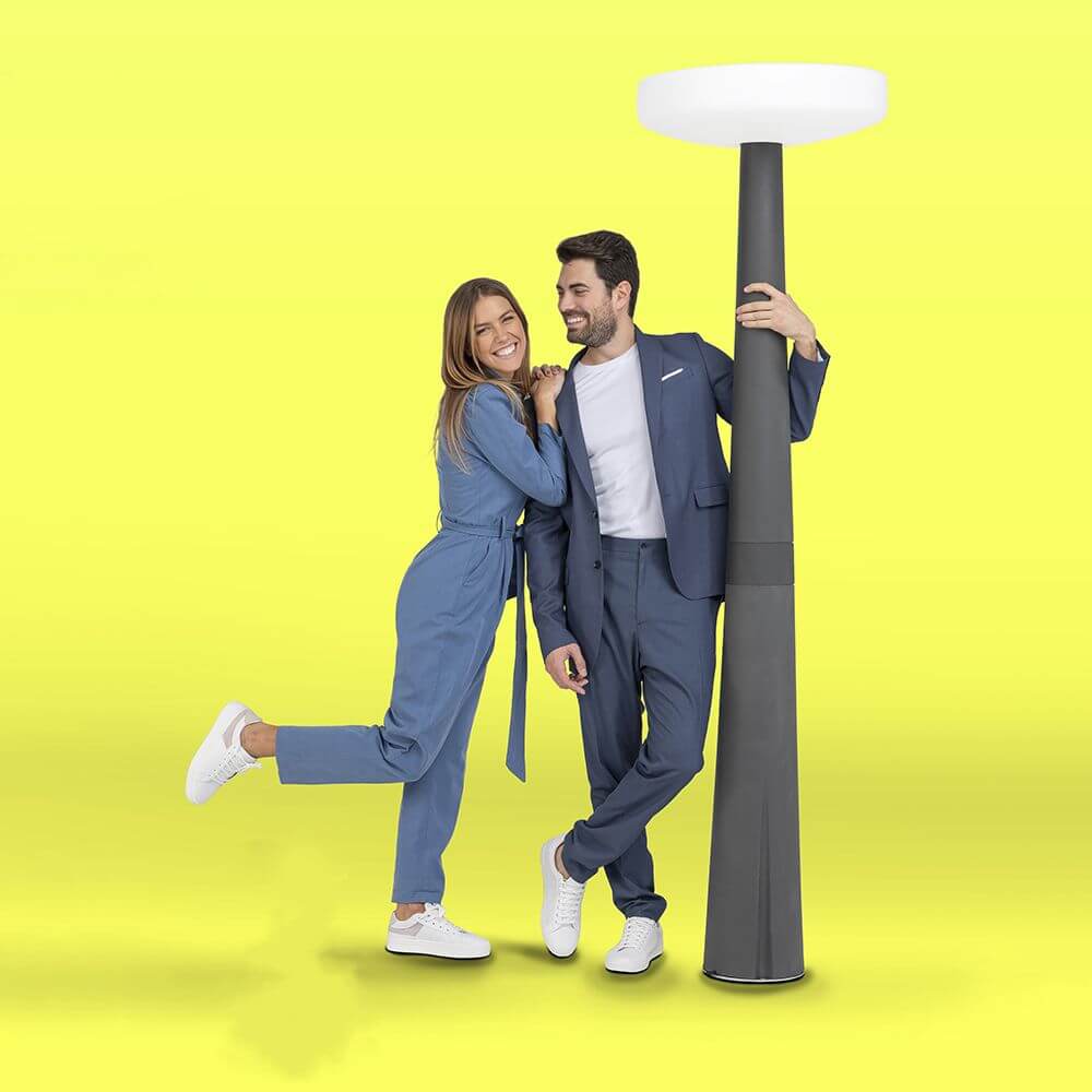 Paar steht lächelnd neben der eleganten LED-Solar-Mastleuchte Paquita Grau vor gelbem Hintergrund.