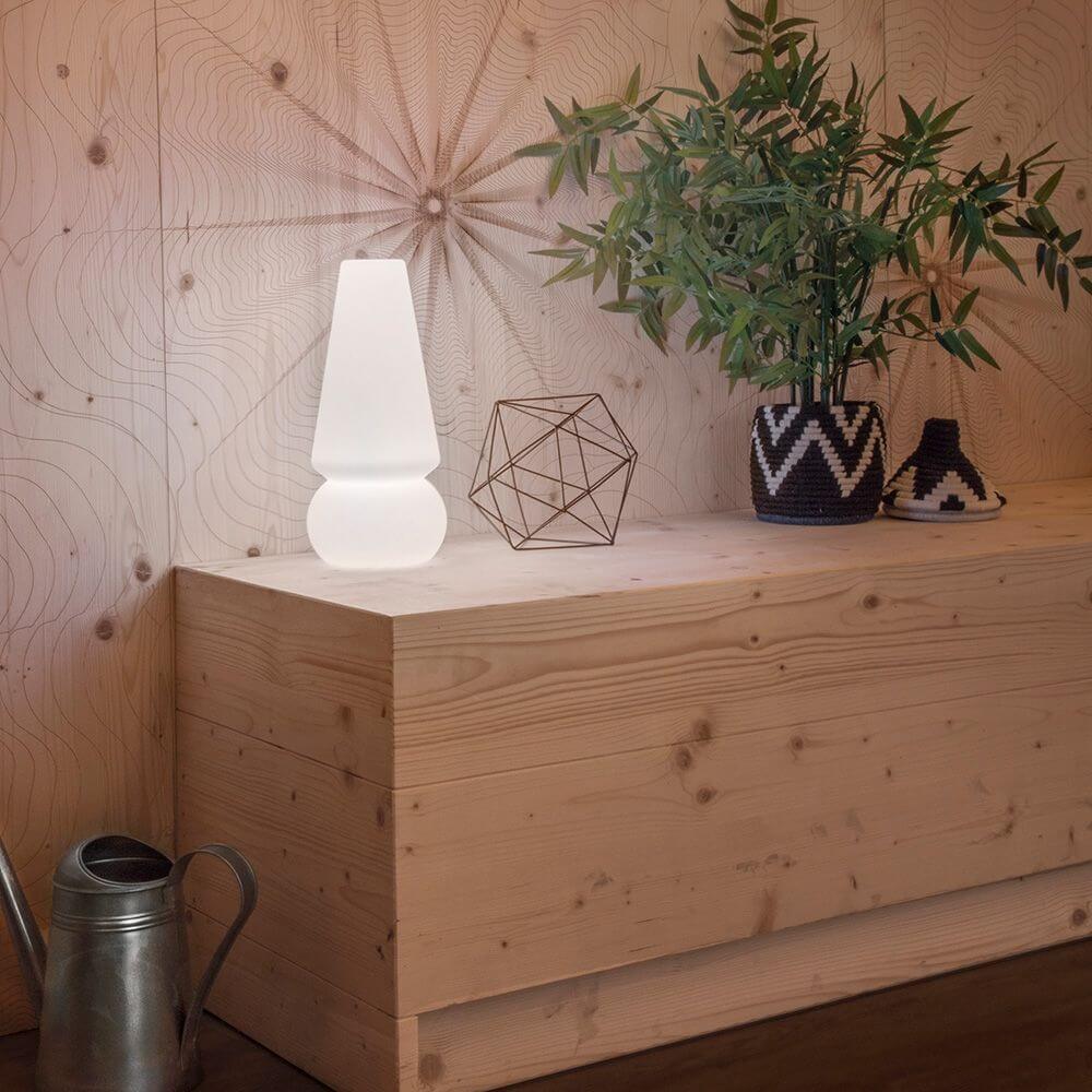 Baby Marge LED-Akkuleuchte auf einem Holzsideboard, umgeben von Pflanzen und Dekoration für eine heitere Atmosphäre.