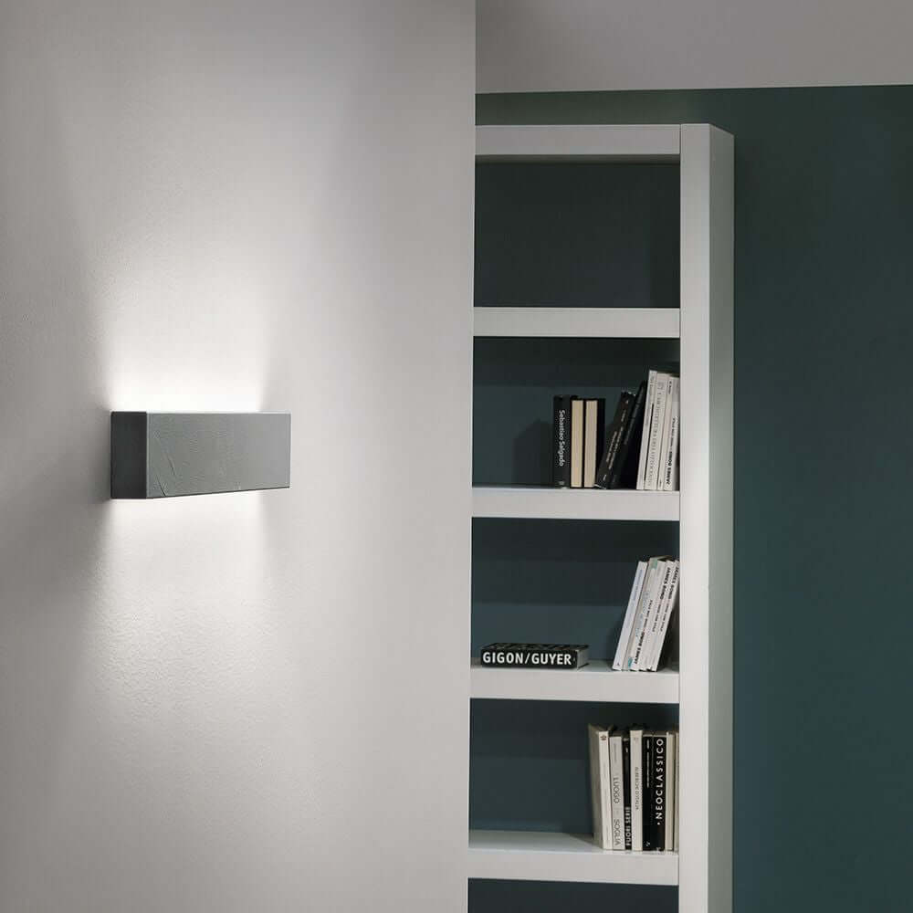 Box W2 LED-Wandleuchte Medium Warmweiß an einer Wand, moderne Einrichtung mit Bücherregal im Hintergrund.