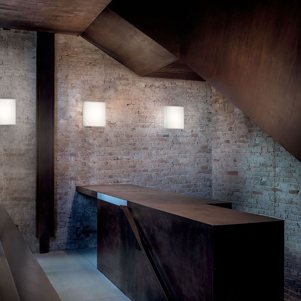 Gluèd W LED-Wandleuchte an einer Ziegelmauer über einem modernen Holzregal.