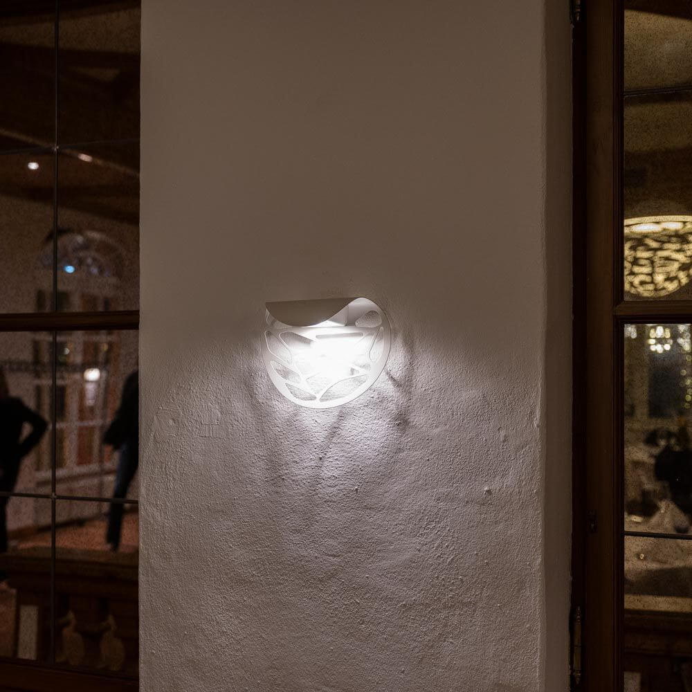 Lodes Kelly Wandlampe an weißer Wand, elegantes Design mit kunstvollem Lichtspiel und Schatteneffekt.