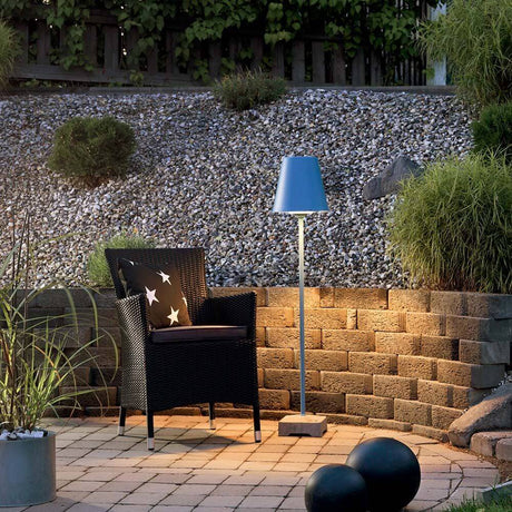 Lucca Terrassenleuchte mit massiven Betonfuß Blau neben einem eleganten Stuhl im Garten.