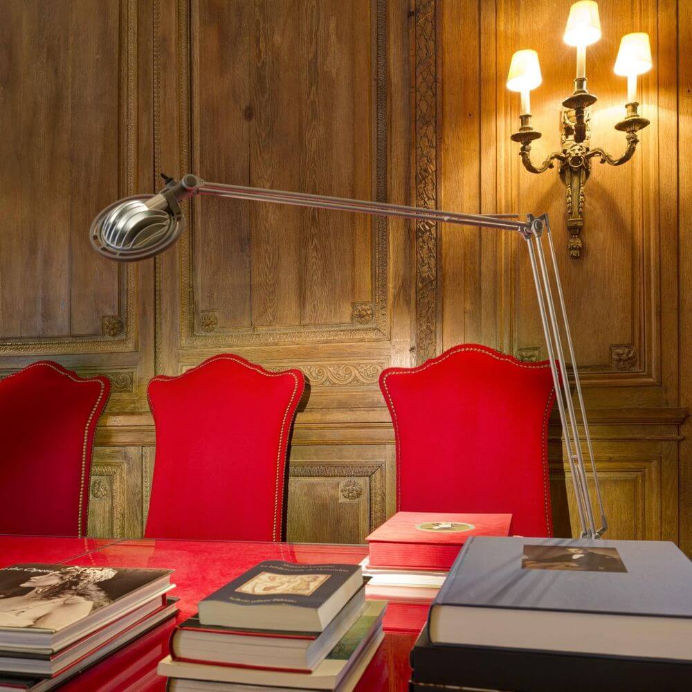 Luceplan LED Schreibtischleuchte Berenice mit Befestigung über einem Tisch mit roten Stühlen und Bücher.