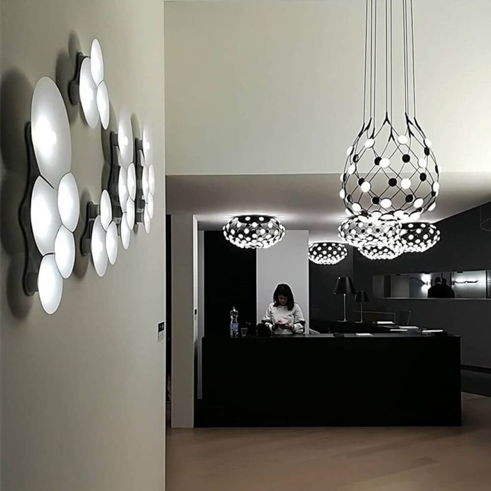 Luceplan LED Wandleuchte Bulbullia 37x31cm an einer modernen Wand, mit stylischen Deckenleuchten im Hintergrund.