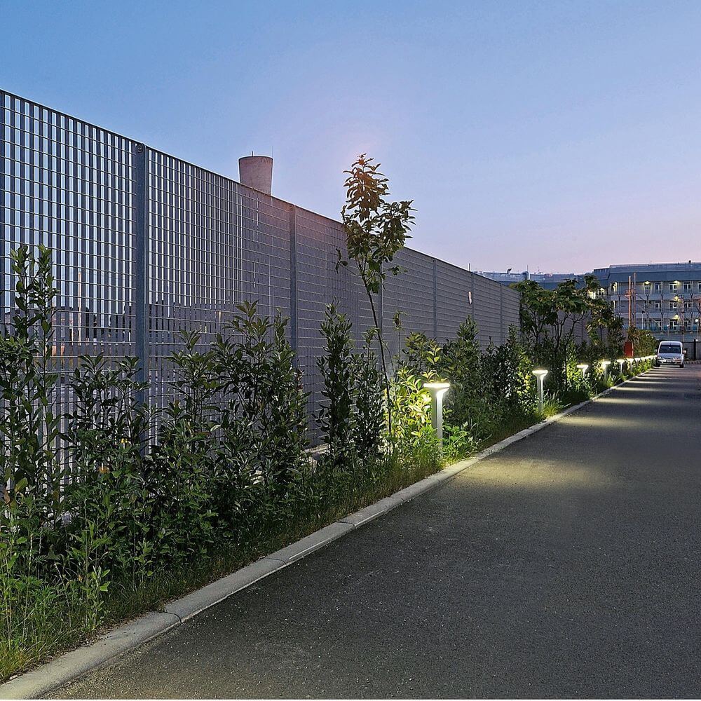 Luceplan Pollerlampe Sky 70cm IP65 beleuchtet einen modernen Weg mit Pflanzen und einem Zaun bei Dämmerung.
