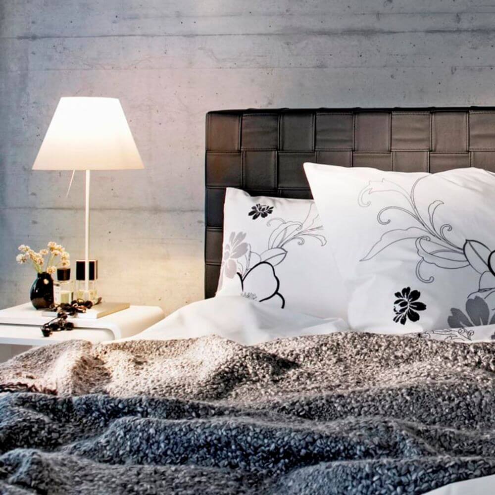 Luceplan Stehlampe Costanza mit Sensor-Dimmer D13 tc neben einem Bett mit floralem Kissen und einer Decke.