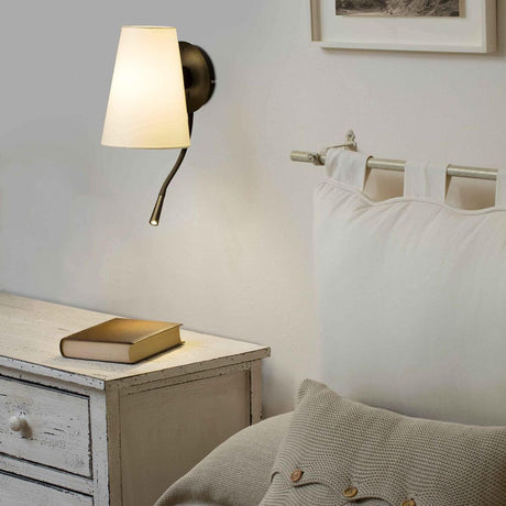 Lupe LED lese-vegglampe svart ved veggen ved siden av en seng med en bok på nattbordet.