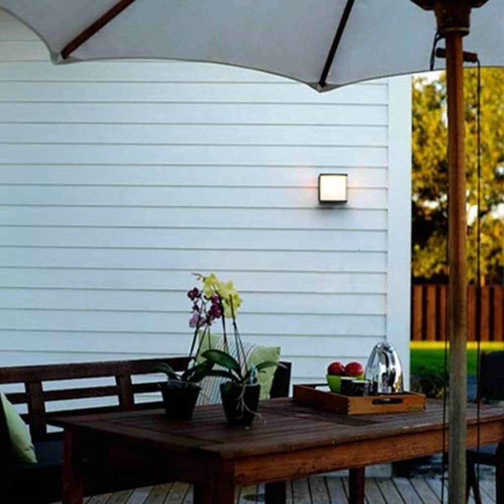 Lutec Außen-Wandleuchte Box Cube IP54 in Anthrazit, an einer Außenwand montiert, mit Tisch und Pflanzen im Vordergrund.