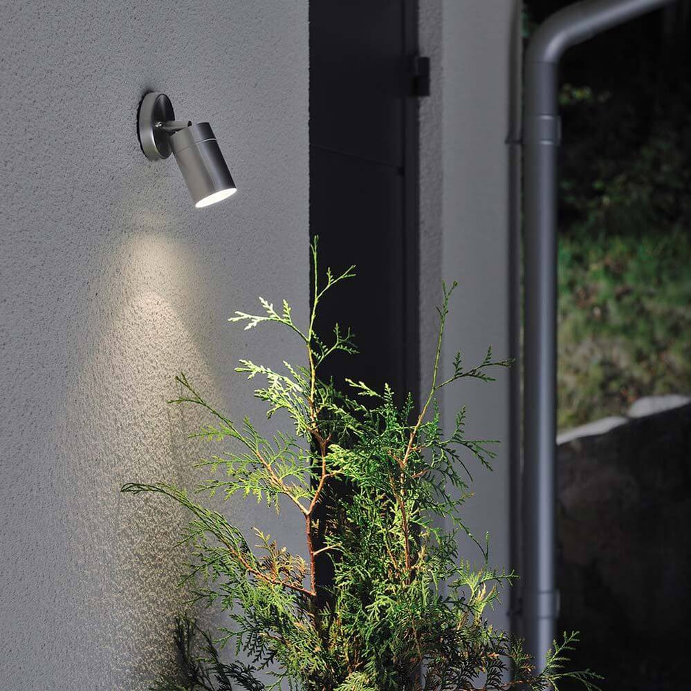 Modena Energiespar Wandspot schwenkbar Edelstahl, klares Glas an einer Wand mit Pflanzen.