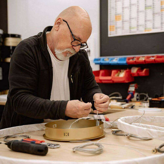 Ein Handwerker arbeitet konzentriert an der Montage eines goldenen Lampenschirms in einer Werkstatt.
