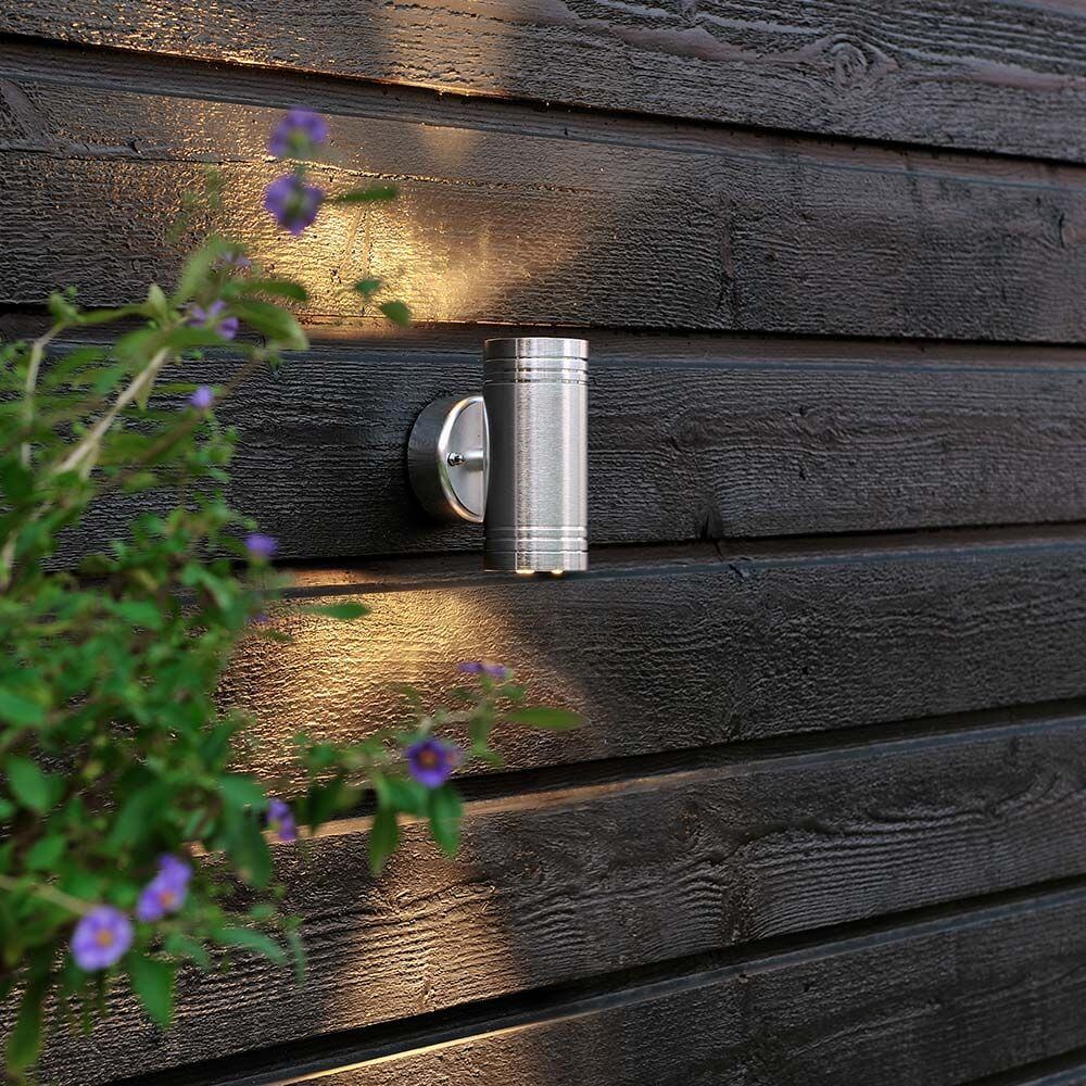 Monza LED lampa ścienna zewnętrzna z podwójnym stożkiem światła na ścianie drewnianej z roślinami na pierwszym planie.