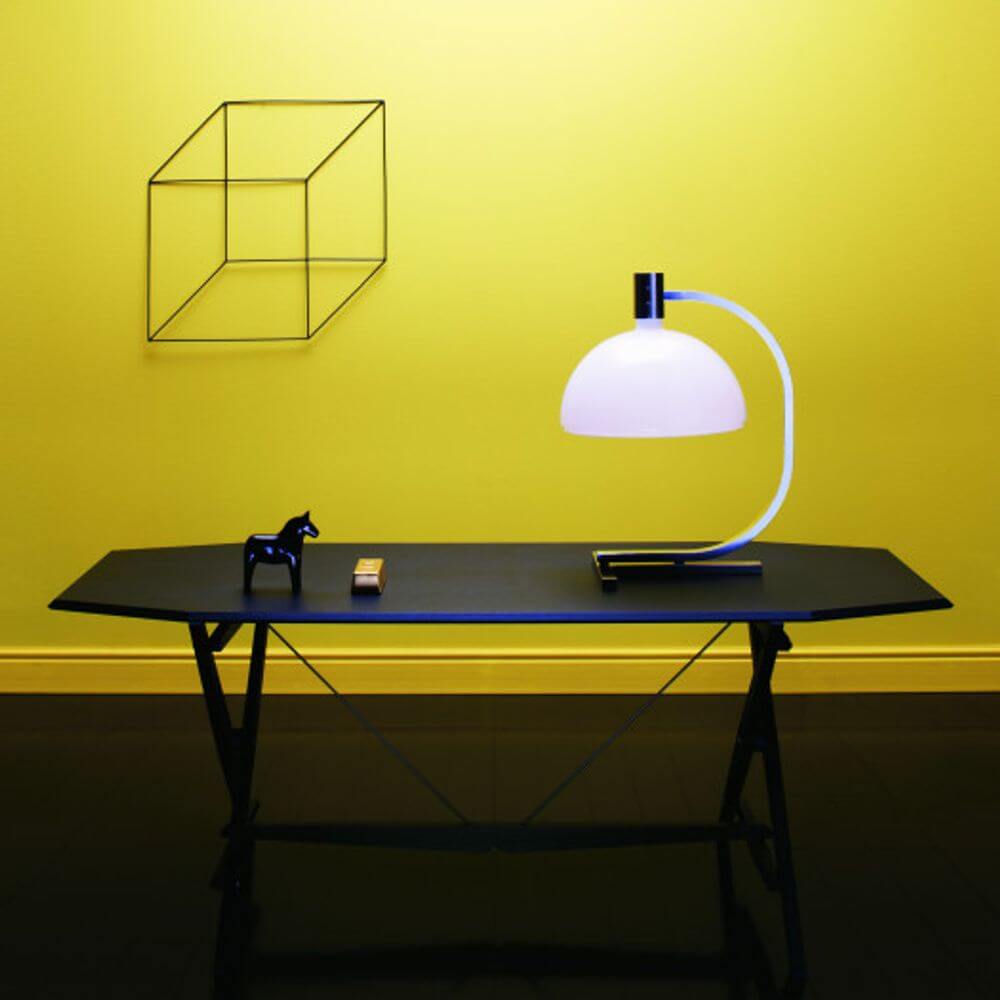 Nemo AS1C Tischlampe Albini mit opalweißem Glasschirm auf schwarzem Tisch vor gelber Wand.