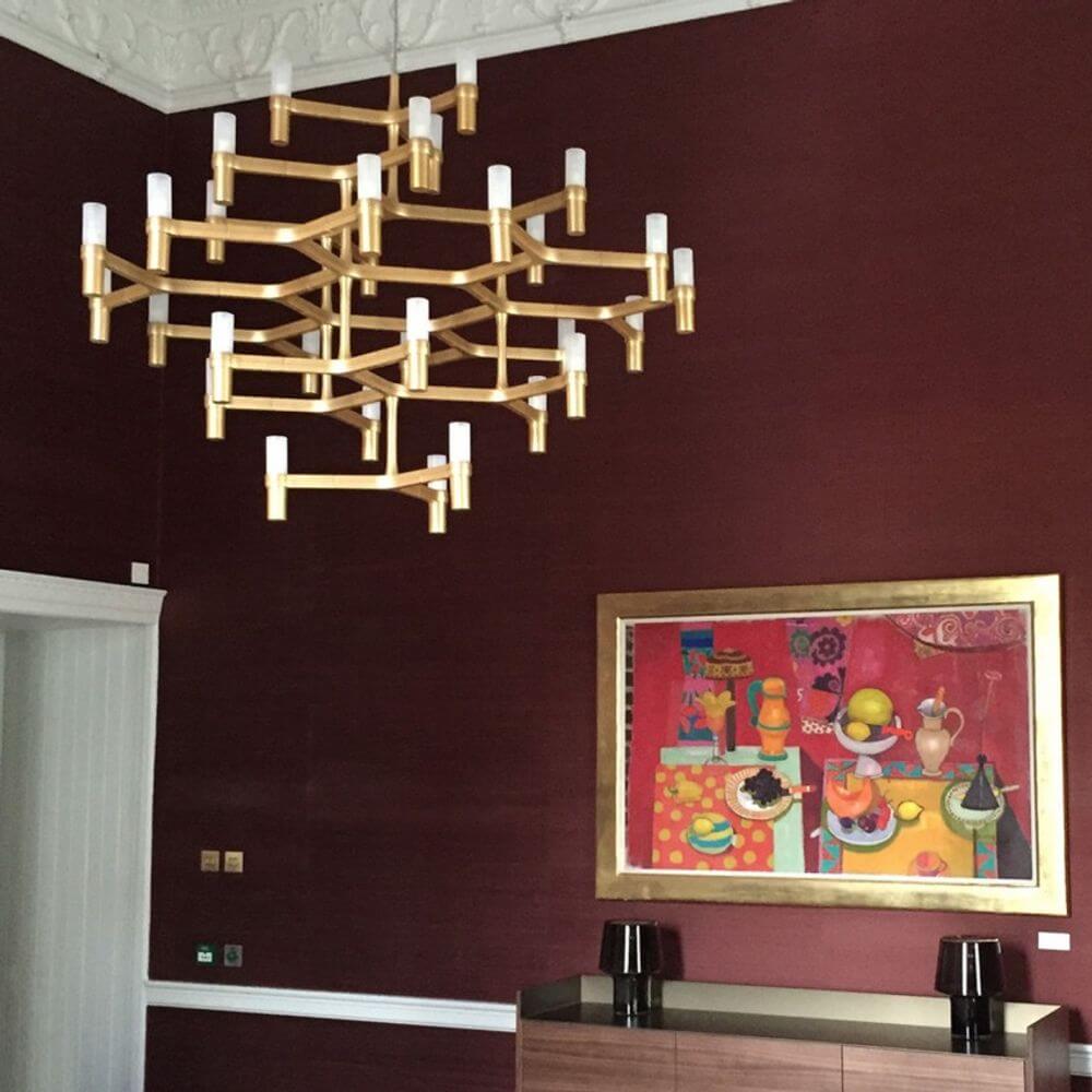 Nemo Crown Major Hängelampe mit 30 Leuchten in gold auf rotem Hintergrund, kombiniert mit farbenfrohem Kunstwerk.