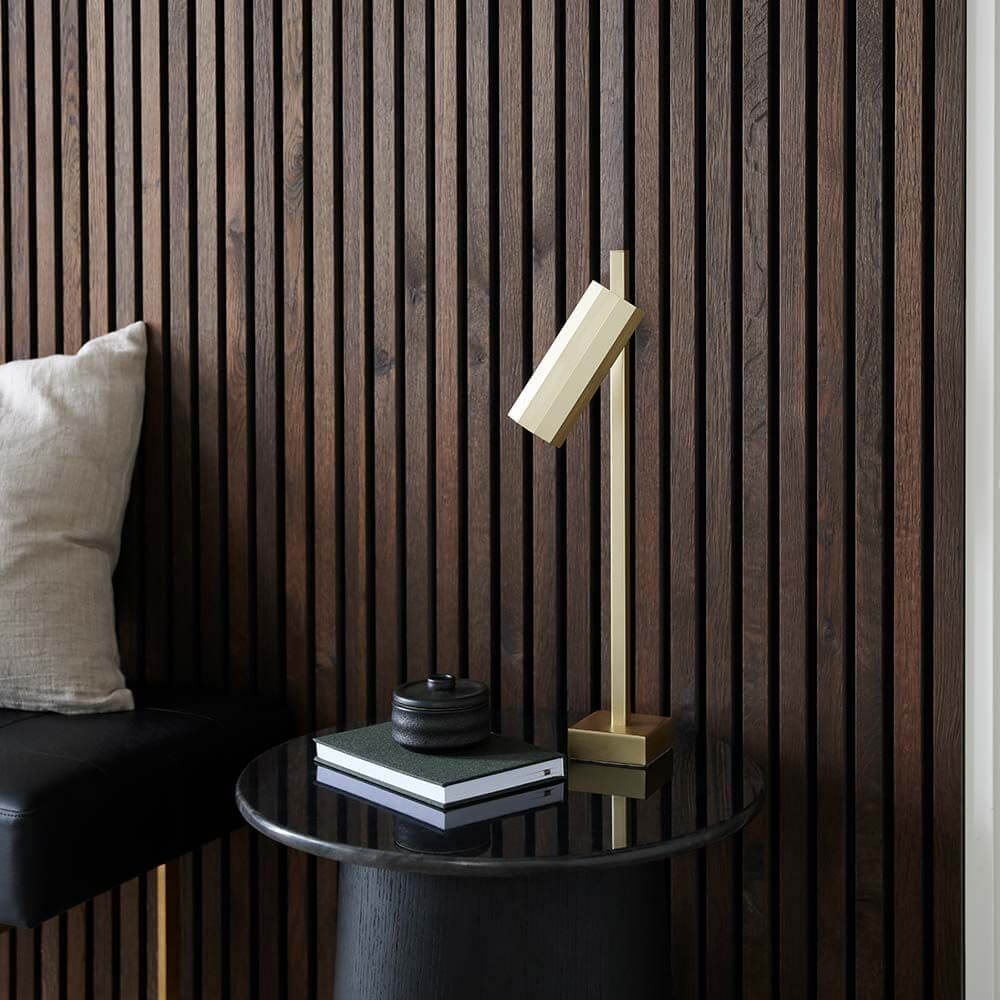 Alanis Tischleuchte auf einem Tisch neben einem Kissen, vor einer Holzvertäfelung mit modernen Linien.