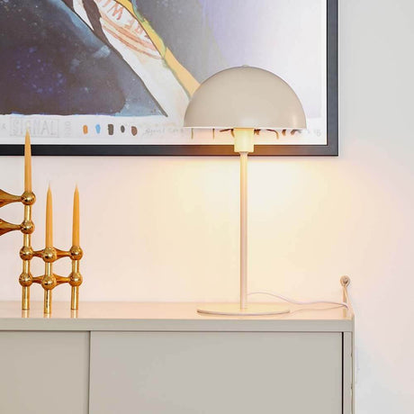 Lampe de table Ellen Beige sur un buffet avec une œuvre d'art moderne en arrière-plan, diffuse une lumière douce.