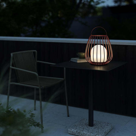 Jim To Go LED rechargeable floor lamp IP54 decoratively placed on a table outdoors, surrounded by a chair and plants.