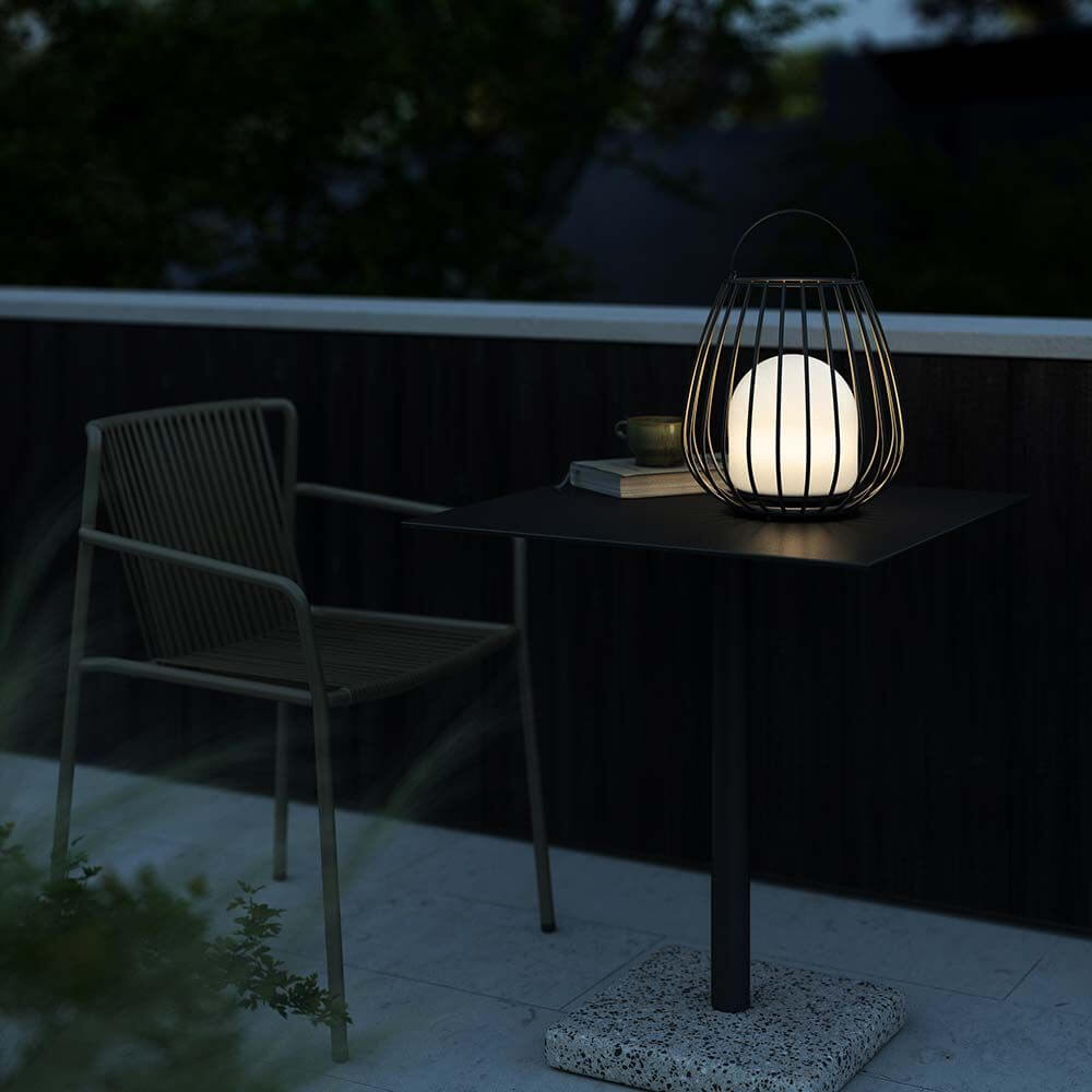 Jim To Go LED Akku Bodenleuchte auf Tisch mit Stuhl im Freien, ideal für Terrasse und stimmungsvolle Beleuchtung.