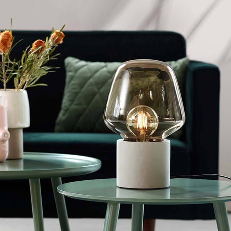 Tischlampe Christina Hellgrau, Rauchfarben auf einem Tisch mit Pflanzen im Hintergrund.