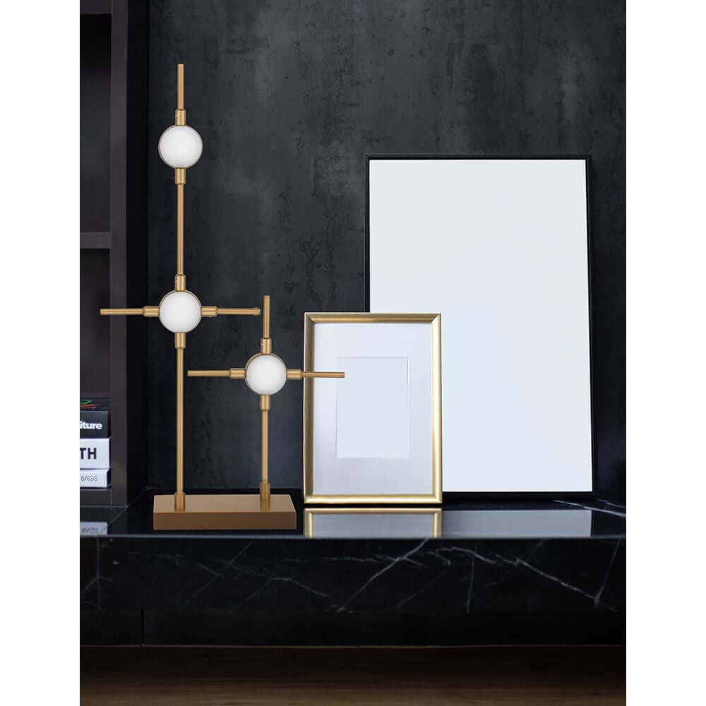 Atomo LED Tischleuchte auf einem modernen Tisch mit goldenen Akzenten und einem leeren Rahmen im Hintergrund.