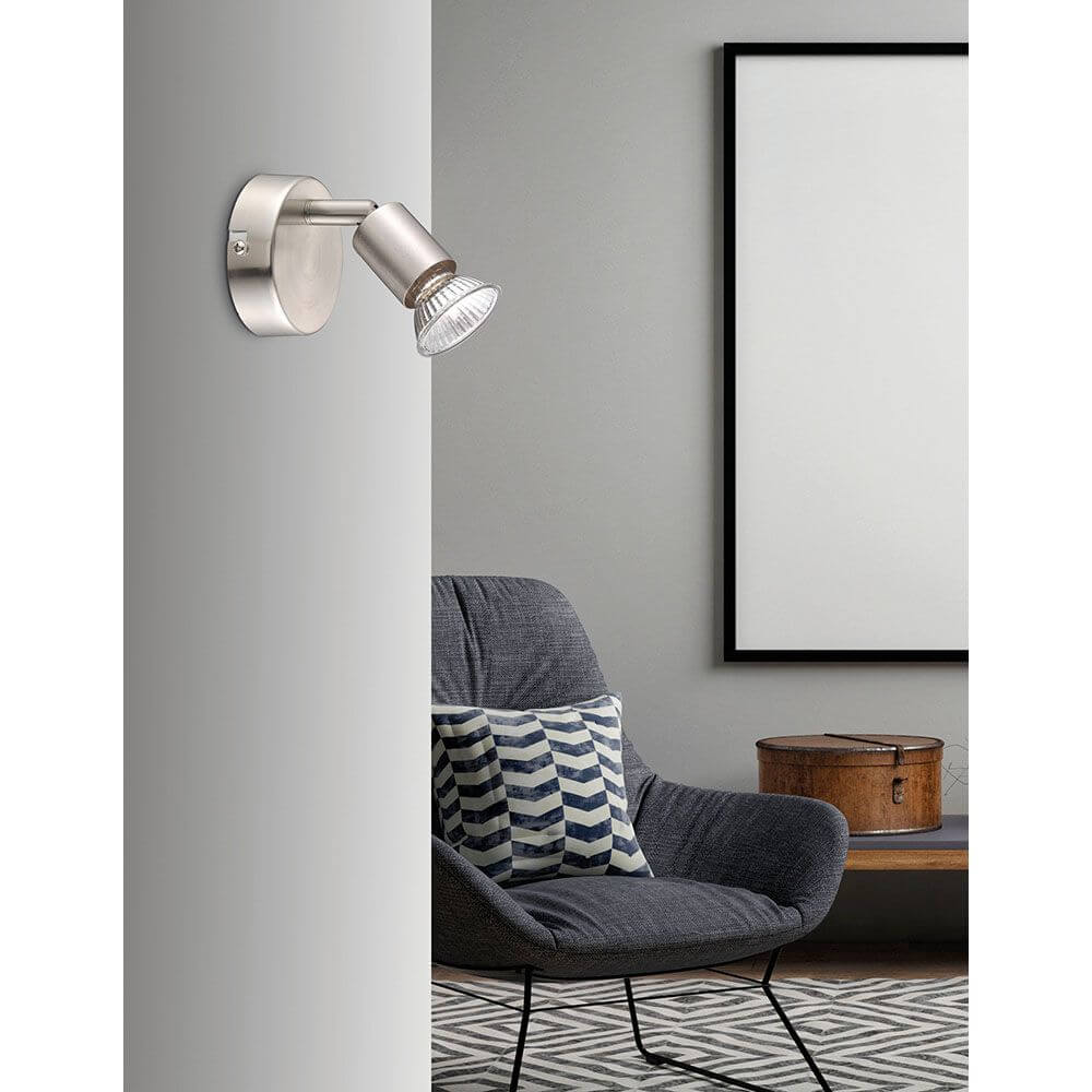 Base Wandspot: moderner Wandstrahler aus Metall mit satiniertem Nickel-Finish über Sessel im Schlafzimmerambiente