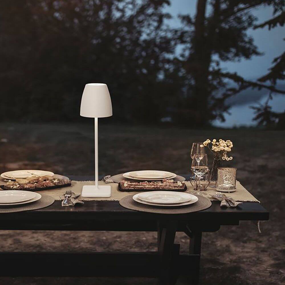 Colt Outdoor-Tischleuchte Akku IP44 Weiß auf einem Tisch im Freien mit gedecktem Geschirr.