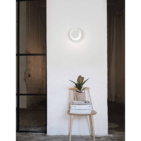 Otto LED wall lamp Ø 12cm white on a wall with a plant on a chair in a modern interior.