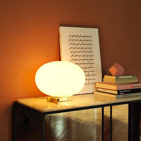 Oluce Glas Tischlampe Alba Alu, Weiß auf einem Tisch mit Bücher und Kunstwerk im Hintergrund.